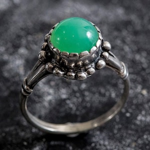 May include: A silver ring with a large, round, bright green gemstone. The gemstone is set in a detailed, ornate bezel. The ring's band has a textured design, and the overall style is vintage or antique.