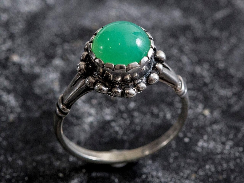 May include: A silver ring with a large, round, bright green gemstone. The gemstone is set in a detailed, ornate bezel. The ring's band has a textured design, and the overall style is vintage or antique.
