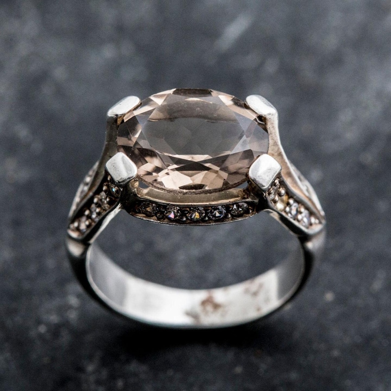 Puede incluir: Un anillo de plata con una gran piedra preciosa ovalada de color marr&oacute;n rodeada de peque&ntilde;as piedras preciosas transparentes.