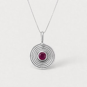 May include: A silver pendant necklace featuring a circular design with concentric grooved rings and a single round, deep pinkish-red gemstone set in the centre. The pendant hangs from a delicate, thin silver chain with a polished metallic finish.