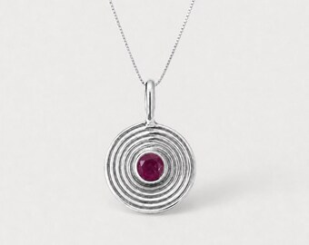 May include: A silver pendant necklace featuring a circular design with concentric grooved rings and a single round, deep pinkish-red gemstone set in the centre. The pendant hangs from a delicate, thin silver chain with a polished metallic finish.