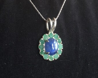 May include: A silver pendant necklace featuring a large oval blue gemstone surrounded by small green gemstones. The pendant hangs from a delicate silver chain, creating a striking contrast against the dark background.
