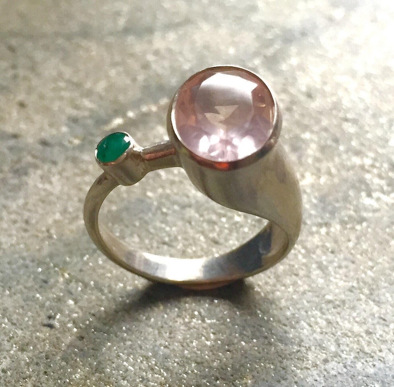 May include: A silver ring with a large pink gemstone and a smaller green gemstone. The ring is set on a textured surface.