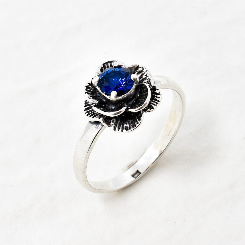 K&ouml;nnte beinhalten: Ein silberner Ring mit einem blauen Edelstein, der in einem Rosenmuster gefasst ist.