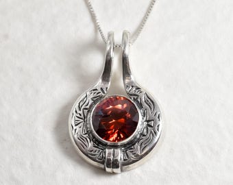 May include: A silver pendant necklace featuring a red gemstone set within an intricate silver frame. The pendant has a distinctive design with two loops at the top.