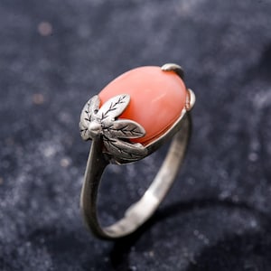 K&ouml;nnte beinhalten: Ein silberner Ring mit einem rosafarbenen ovalen Edelstein, der in einem floralen Design gefasst ist.