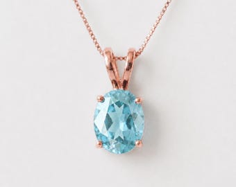 Puede incluir: Un collar de oro rosa con un colgante de piedra preciosa azul claro de corte ovalado. La piedra facetada se sujeta mediante un engaste de cuatro garras unido a una delicada cadena de oro rosa. El diseño resalta el contraste entre el metal cálido y el azul frío.