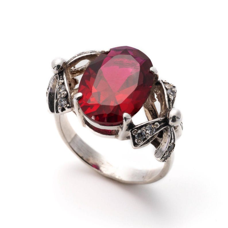 May include: A silver ring with a large, oval-shaped red gemstone surrounded by smaller clear gemstones. The ring has a delicate, ornate design.