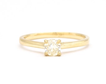 May include: A gold solitaire ring with a round, clear gemstone. The ring has a thin band and a four-prong setting, showcasing the central stone. The ring is set against a white background.