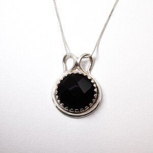 Puede incluir: Un collar con colgante de plata que presenta una piedra negra redonda y facetada, engastada en un marco de plata pulida con un asa decorativa en forma de bucle. El colgante cuelga de una cadena de plata fina con un acabado met&aacute;lico brillante.