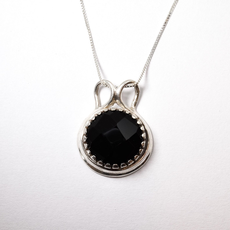 Puede incluir: Un collar con colgante de plata que presenta una piedra negra redonda y facetada, engastada en un marco de plata pulida con un asa decorativa en forma de bucle. El colgante cuelga de una cadena de plata fina con un acabado met&aacute;lico brillante.