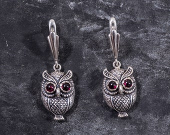 May include: A pair of silver owl earrings with red gemstone eyes. The owls are detailed with textured feathers and have a dangling design.