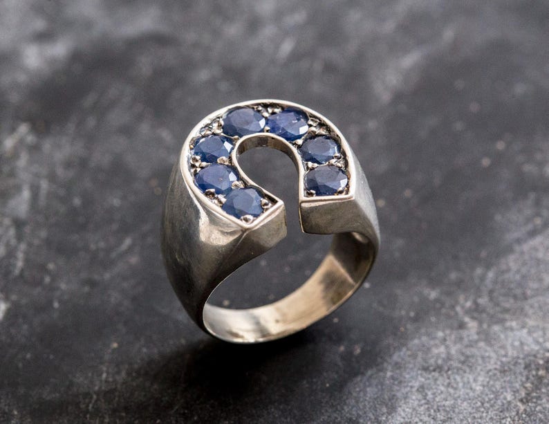K&ouml;nnte beinhalten: Ein silberner Ring mit einem Hufeisen-Design, besetzt mit blauen Edelsteinen.