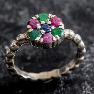 May include: A silver ring with a floral design. The ring features a cluster of gemstones in shades of green, pink, and blue. The band has a textured, beaded design. The ring is set against a dark background.