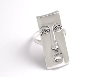 May include: A silver ring with a face design. The face is sculpted in a rectangular shape and has a serious expression.