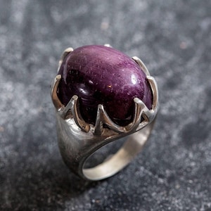 May include: A silver ring featuring a large, oval-shaped, deep purple gemstone. The gemstone is set in a crown-like silver setting. The ring's band is smooth and polished, creating a striking contrast with the textured gemstone.