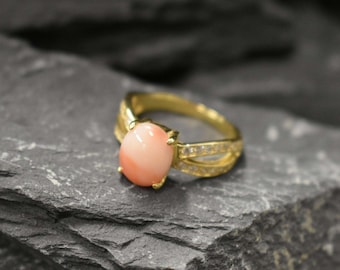 Può includere: Un anello d'oro con una pietra preziosa di corallo rosa e un delicato design a croce. L'anello è incastonato con piccole pietre trasparenti.