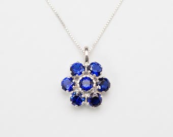 Pode incluir: Um colar de prata com um pendente em forma de flor, cravejado com sete pedras redondas facetadas de azul profundo. O pendente está suspenso numa delicada corrente de prata, com as pedras dispostas num grupo que lembra uma flor aberta.