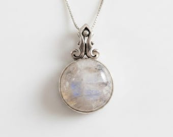 May include: A silver pendant necklace featuring a round, iridescent moonstone. The moonstone is set in a decorative silver bezel and hangs from a delicate silver chain. The pendant has an ornate, stylized bail.