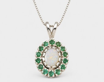 May include: A silver pendant necklace featuring a central oval-shaped iridescent opal surrounded by a halo of small, round, vibrant green emerald gemstones. The pendant hangs from a delicate silver chain with a classic box link design.