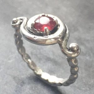 May include: A silver ring with a red gemstone set in a swirl design. The band is textured and has a delicate, intricate design.