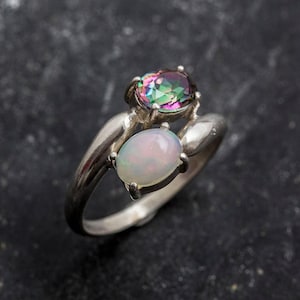 Può includere: Un anello d'argento con due pietre ovali. La pietra più grande è un opale arcobaleno, e la pietra più piccola è un opale bianco.