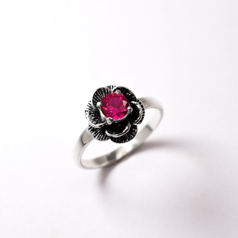 Może przedstawiać: Srebrny pierścionek z r&oacute;żowym kamieniem szlachetnym osadzonym w kwiatowym wzorze.