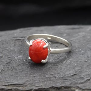May include: A silver ring with a vibrant red oval gemstone. The ring's band is a polished silver, and the gemstone is securely held in place by four prongs. The ring is set against a dark, textured background.