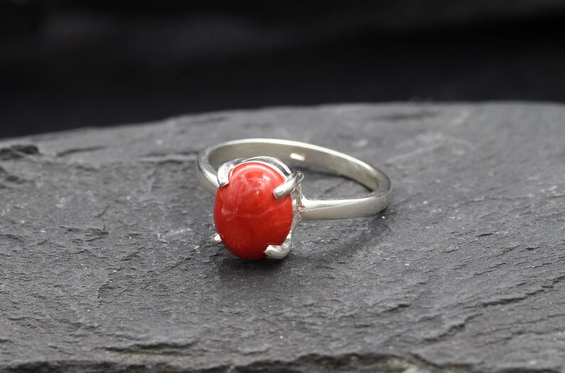 May include: A silver ring with a vibrant red oval gemstone. The ring's band is a polished silver, and the gemstone is securely held in place by four prongs. The ring is set against a dark, textured background.