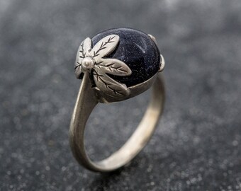 Könnte beinhalten: Ein silberner Ring mit einem dunkelblauen, gesprenkelten Edelstein. Der Edelstein ist in einer floralen Lünette gefasst. Das Band des Rings ist aus poliertem Silber. Das Design ist elegant und alltagstauglich.
