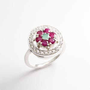 Op de afbeelding: Een zilveren ring met een rond middenstuk met een cluster van roze en groene edelstenen, omringd door kleinere witte edelstenen.