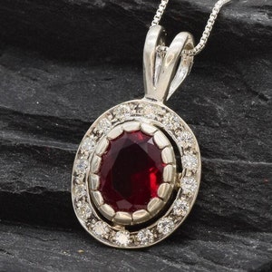 Pode incluir: Um colar com um pingente de prata com uma pedra oval vermelha cercada por um halo de pedras brancas menores.