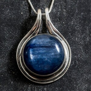 May include: A silver pendant with a round blue gemstone set in a silver frame. The pendant is on a silver chain.