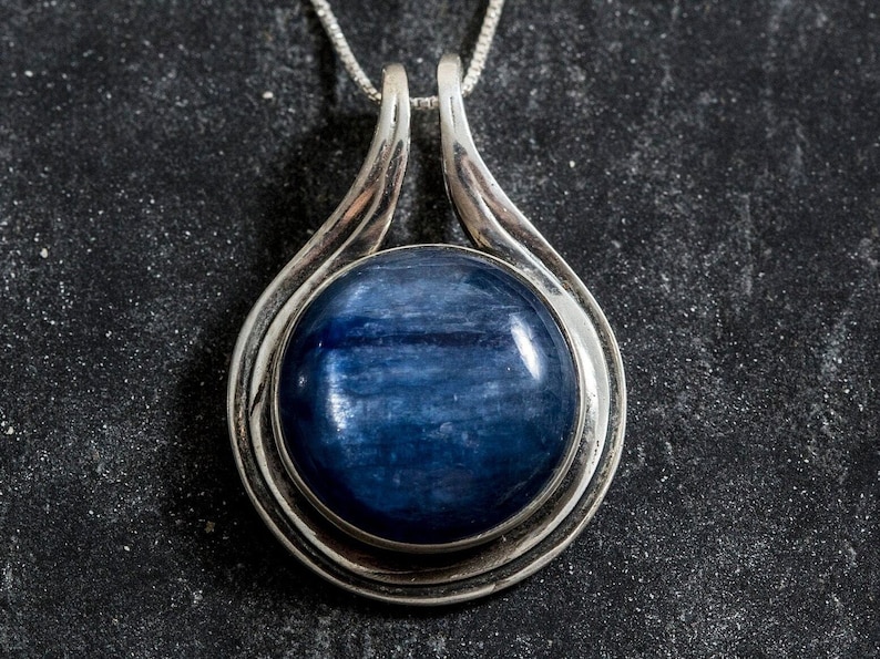 May include: A silver pendant with a round blue gemstone set in a silver frame. The pendant is on a silver chain.