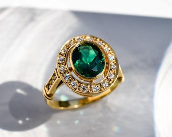 May include: A gold ring featuring a large oval-cut green gemstone at the center, surrounded by a halo of small clear stones. The band is made of polished gold and includes additional small clear stones set along the sides of the ring shank.