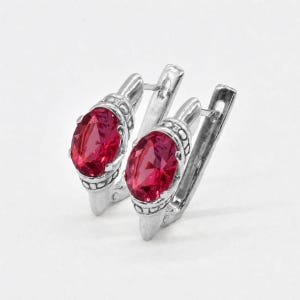 May include: A pair of silver-toned earrings featuring oval-cut, vibrant red faceted stones. Each earring is set in a polished metal frame with decorative beaded detailing around the base of the stone and a secure hinged lever-back closure.