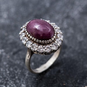 May include: A silver ring featuring a large oval purple gemstone, encircled by a halo of smaller clear gemstones.