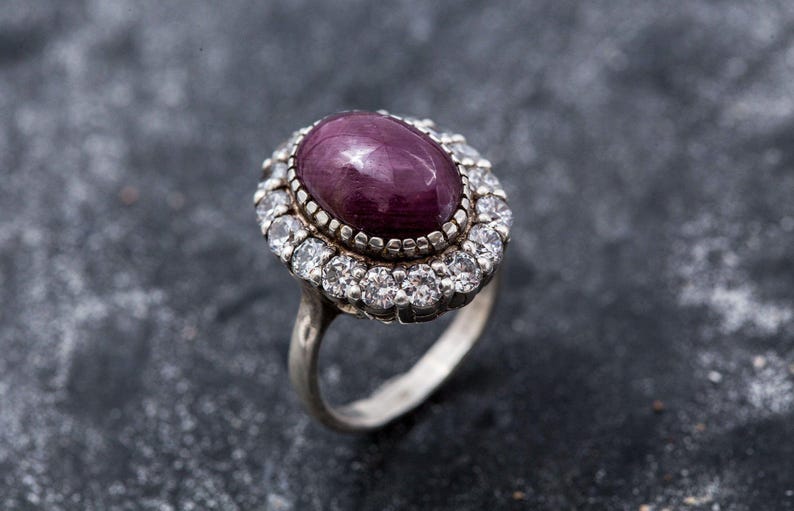 May include: A silver ring featuring a large oval purple gemstone, encircled by a halo of smaller clear gemstones.