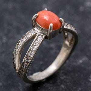 Pu&ograve; includere: Un anello d'argento con una pietra preziosa di corallo e un design a croce, accentuato da piccole pietre chiare.