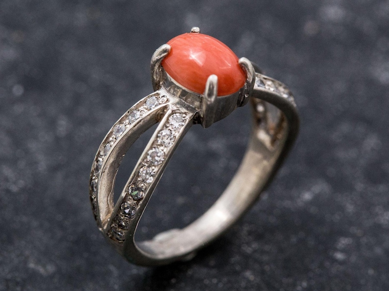 Pu&ograve; includere: Un anello d'argento con una pietra preziosa di corallo e un design a croce, accentuato da piccole pietre chiare.