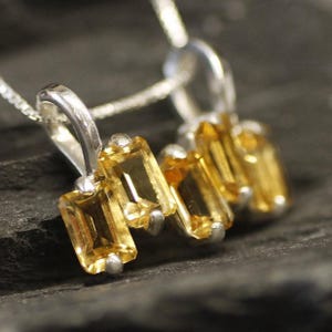 May include: A silver necklace with a pendant featuring five rectangular, faceted, topaz gemstones in a graduated size. The gemstones are a warm, golden yellow color.