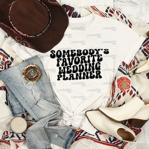 May include: A white t-shirt with the text "SOMEBODY'S FAVORITE WEDDING PLANNER" in black. The shirt is styled with blue jeans, a brown cowboy hat, a bracelet, and white ankle boots. A decorative flower and a patterned blanket complete the ensemble.