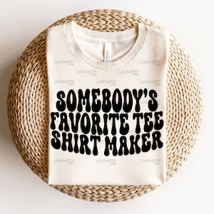 May include: A white t-shirt with the text "Somebody's Favorite Tee Shirt Maker" in black, retro-style lettering.