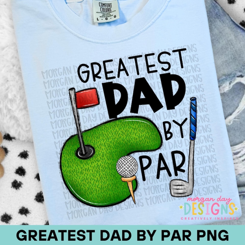May include: Pale blue t-shirt with a golf-themed design. The graphic includes the words "GREATEST DAD" in black, a green golf course, a golf ball, and a golf club. The phrase "BY PAR" is also part of the design.
