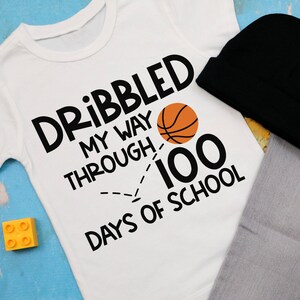 Puede incluir: Camiseta blanca con texto negro que dice "Dribbled my way through 100 days of school" con un gr&aacute;fico de baloncesto y una l&iacute;nea discontinua.