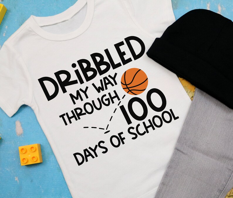 Puede incluir: Camiseta blanca con texto negro que dice "Dribbled my way through 100 days of school" con un gr&aacute;fico de baloncesto y una l&iacute;nea discontinua.