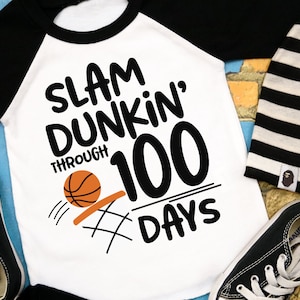 May include: A white and black baseball-themed shirt with the text "Slam Dunking Through 100 Days" and a basketball graphic.