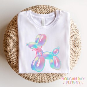 May include: White t-shirt with a colorful balloon animal design. The balloon animal is a dog with iridescent colors and a pink ribbon.