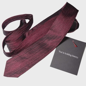 May include: A burgundy tie with a subtle dotted pattern and a matching tag. The tag reads "You're holding history." The tie is made of a textured fabric and is suitable for formal occasions.