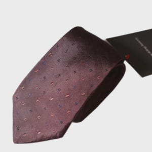May include: A burgundy tie with a repeating pattern of small, evenly spaced rectangles in shades of blue and red. The tie is made of a smooth, possibly silk, material and is neatly folded. A black tag is attached.